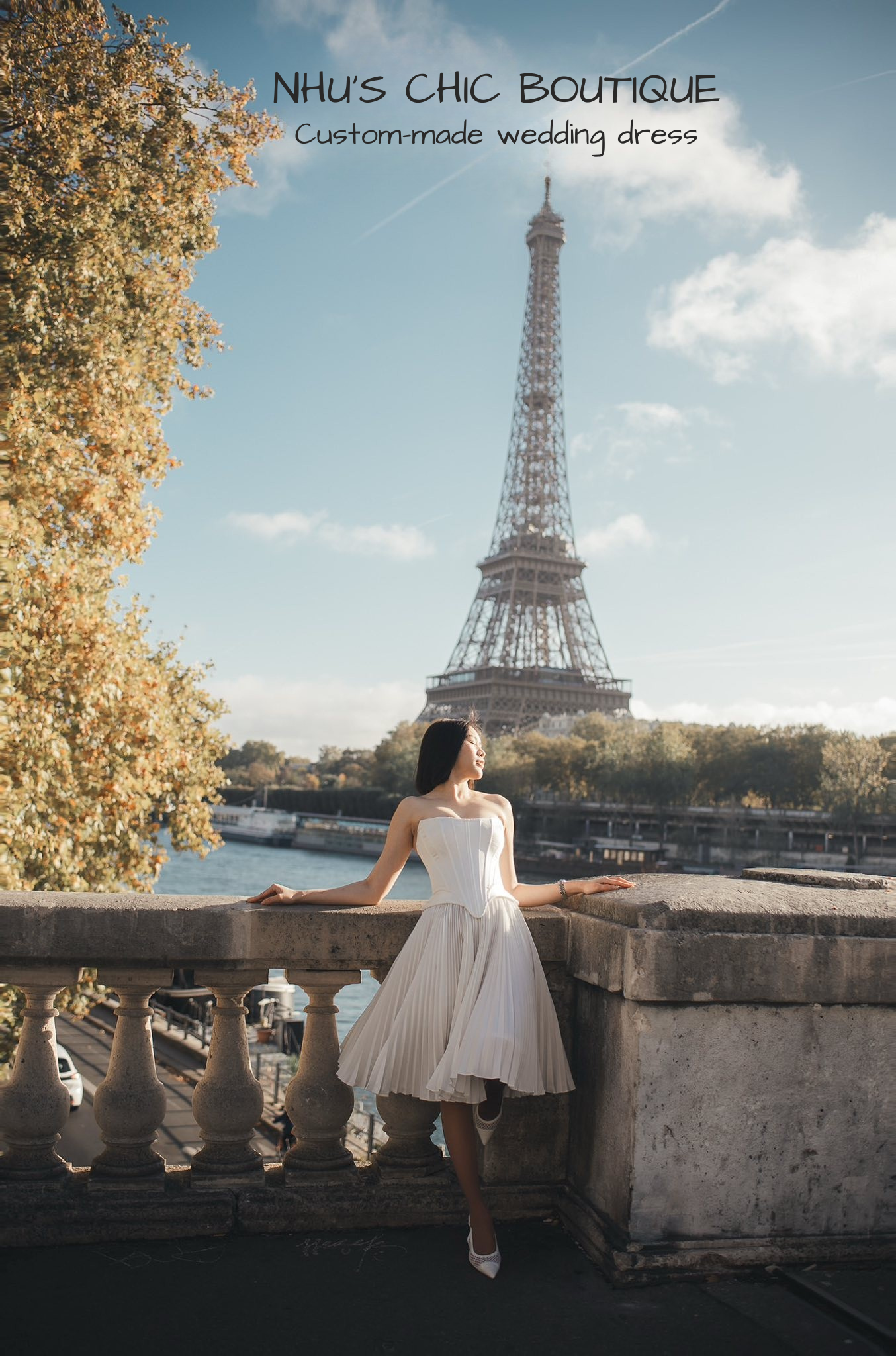 A bride wearing the Genevieve pleated tea-length wedding dress leaning against a stone railing with the Eiffel Tower in the background.