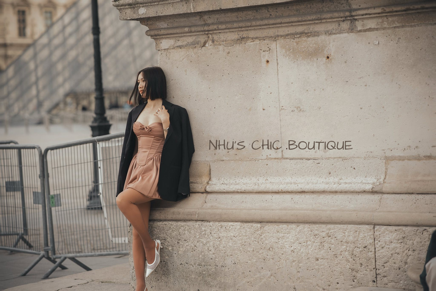 A woman wearing the Chloe caramel corset mini dress paired with a black blazer, leaning against a stone wall near the Louvre Museum in Paris.