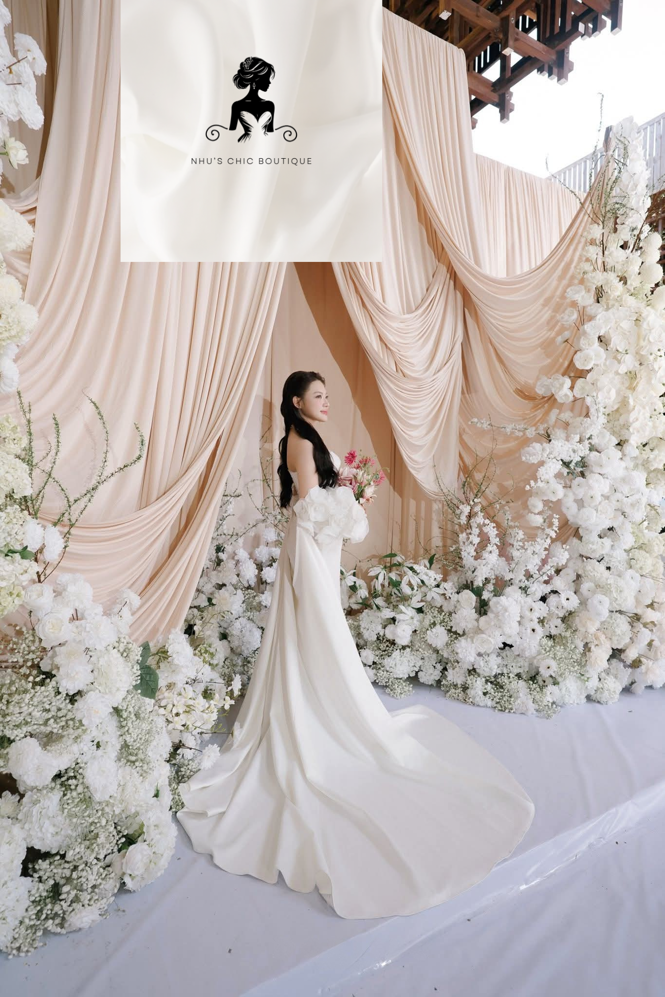 Full-length side profile of a bride in a white mermaid-style wedding dress with a long, elegant train. She is standing in front of a grand floral installation of white hydrangeas and roses, framed by draped fabric. The image features the Nhu’s Chic Boutique logo.