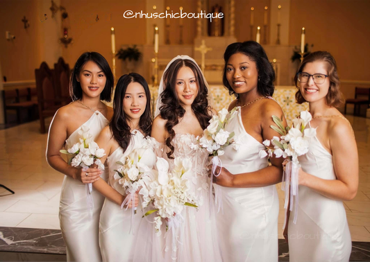 Bride wearing the Alice Dress standing with bridesmaids in sleek white halter-neck gowns.