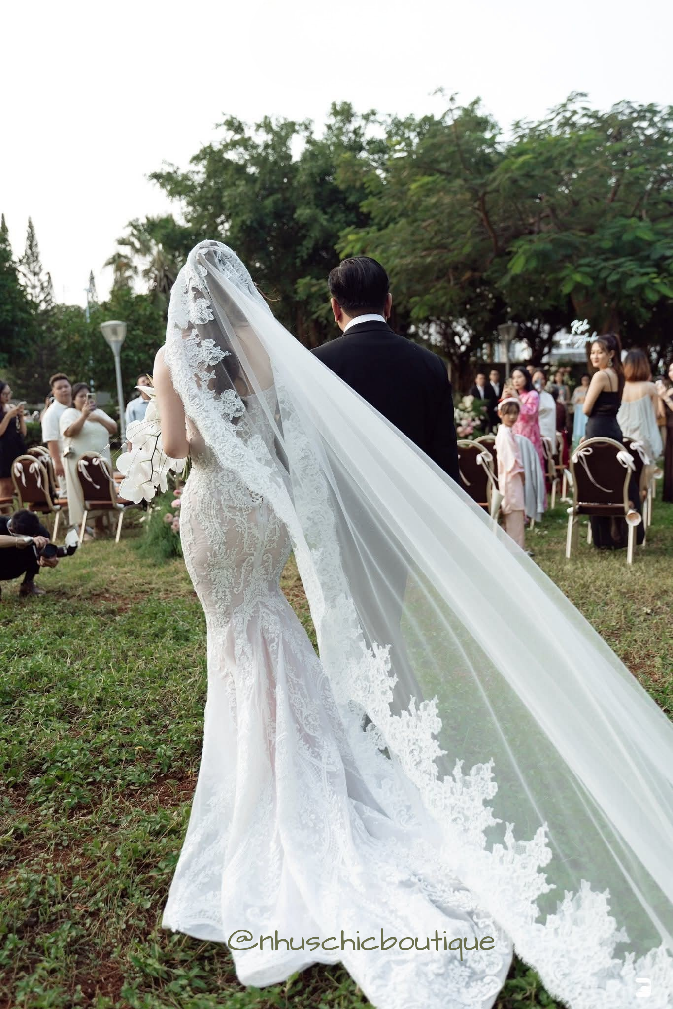 Back view of a bride in a mermaid lace wedding dress with a long cathedral-length lace-edged veil walking through a garden.