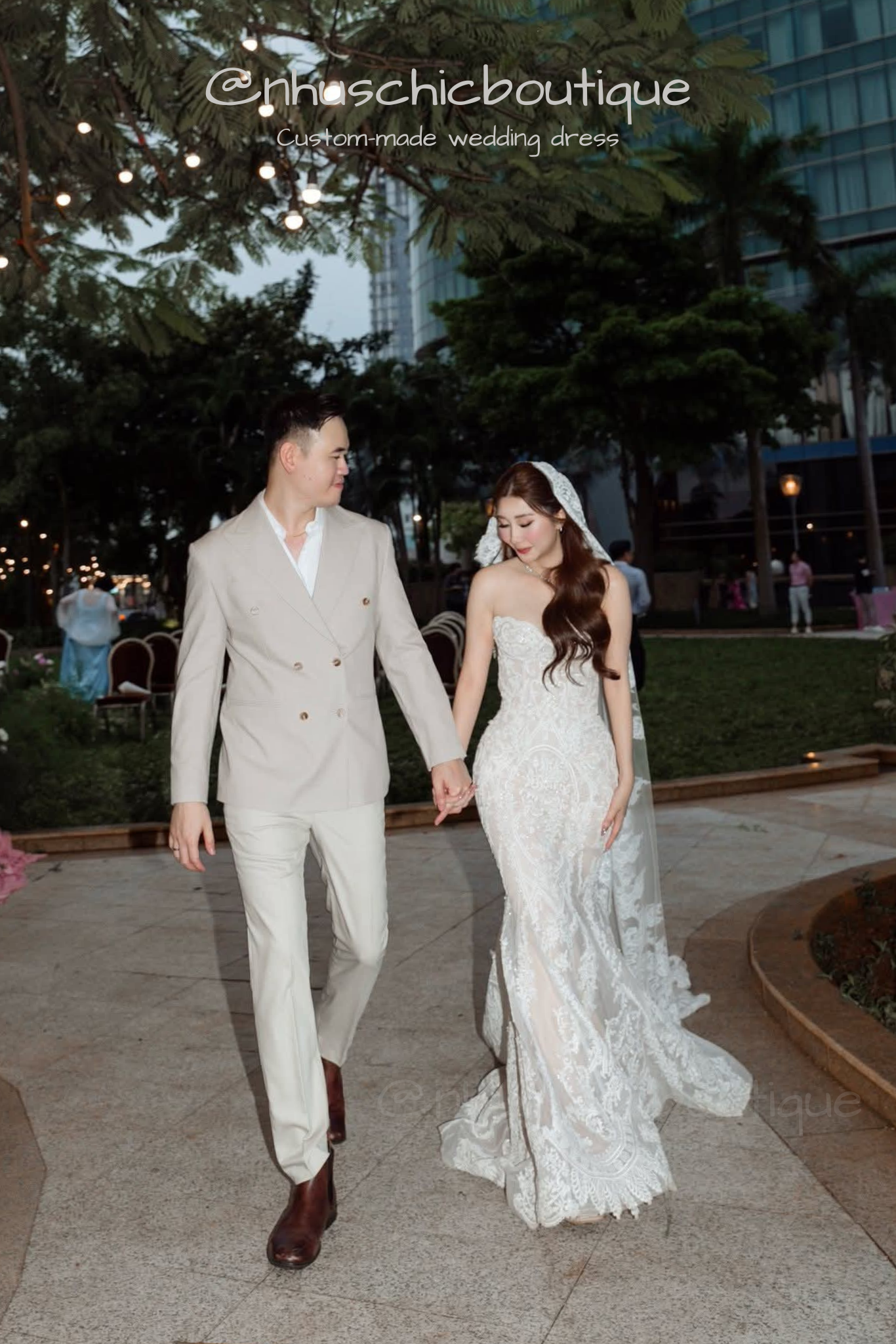 Couple holding hands; bride in a strapless sweetheart lace mermaid gown and groom in a beige double-breasted suit.