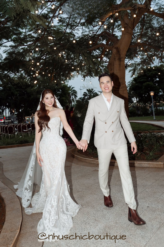 Couple holding hands; bride in a strapless sweetheart lace mermaid gown and groom in a beige double-breasted suit.