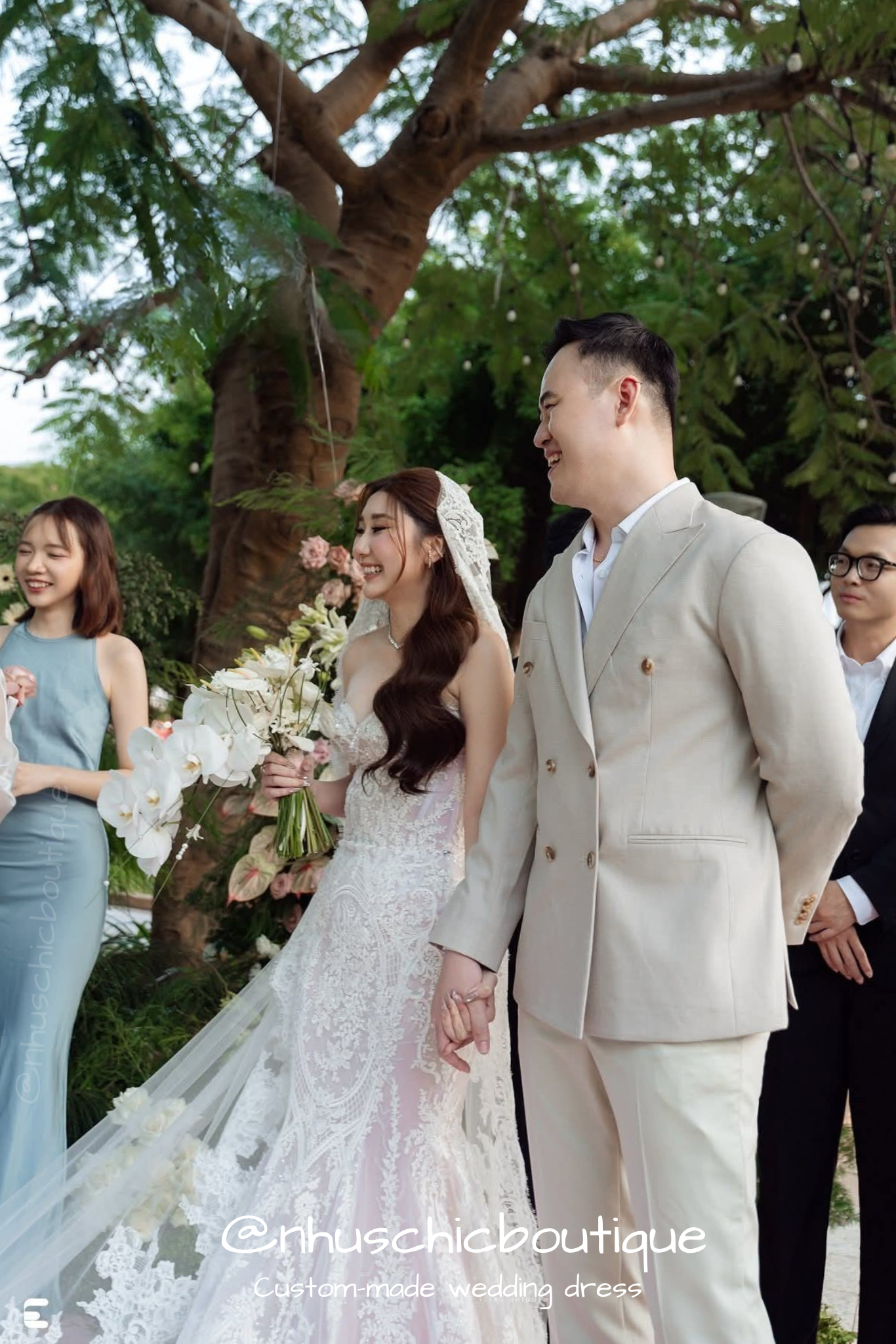 Couple holding hands; bride in a strapless sweetheart lace mermaid gown and groom in a beige double-breasted suit.
