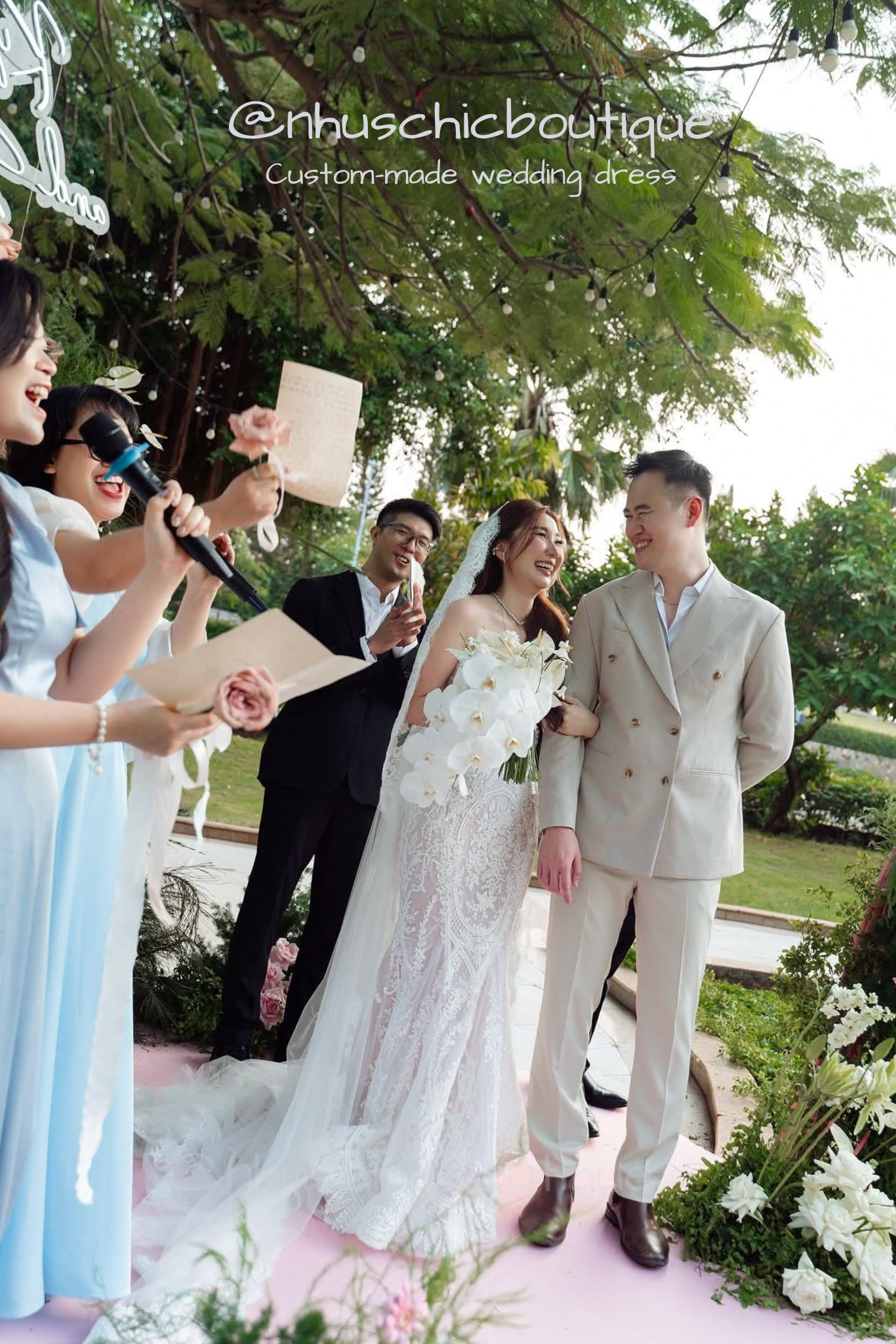Wedding couple standing outdoors with guests, surrounded by greenery.